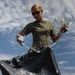 A person wearing military-style clothing is engaged in a cleanup activity, collecting debris into a large black trash bag. They are wearing tactical gloves and sunglasses while holding what appears to be plastic strapping or ribbon-like waste material. The photo is taken from a low angle against a partly cloudy sky, giving a dramatic perspective.