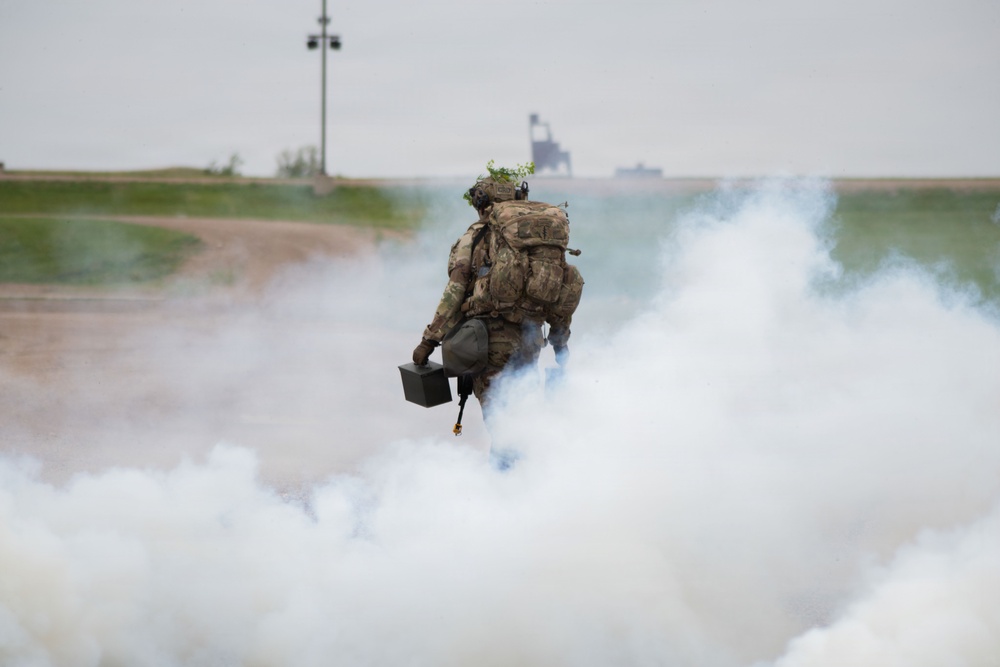 A soldier in full combat gear and camouflage uniform is moving through a dense cloud of white smoke, viewed from behind. The individual is carrying a large rucksack and what appears to be an ammunition can in one hand. The scene appears to be a military training exercise conducted near a waterway or port area.