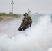 A soldier in full combat gear and camouflage uniform is moving through a dense cloud of white smoke, viewed from behind. The individual is carrying a large rucksack and what appears to be an ammunition can in one hand. The scene appears to be a military training exercise conducted near a waterway or port area.