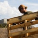 A person wearing an olive green shirt, sunglasses, and black work gloves is lifting or moving a large wooden shipping pallet outdoors. The setting appears to be a dry, grassy field or training area with a structure visible in the background. The image is taken from a low angle, emphasizing the scale of the pallet relative to the individual.