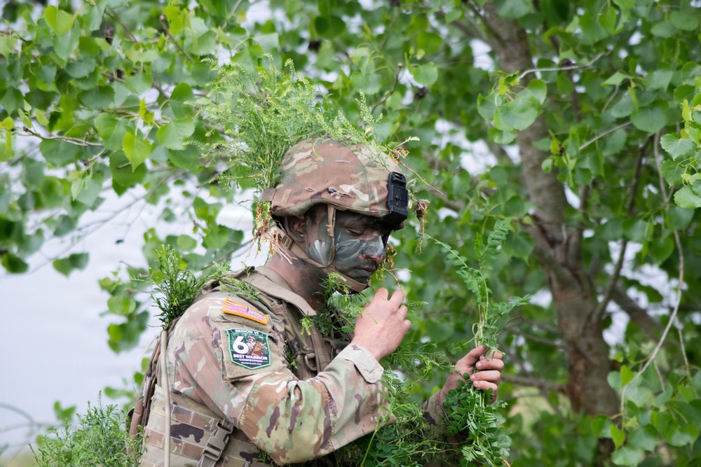 A U.S. Army soldier in full camouflage gear, including face paint and vegetation attached to their helmet and uniform, is engaged in a concealment or stalking exercise. The soldier wears a patch identifying them as a participant in the 'Region VI Best Warrior' competition. They are holding foliage and appear to be in the process of applying or arranging natural camouflage material.
