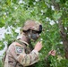 A U.S. Army soldier in full camouflage gear, including face paint and vegetation attached to their helmet and uniform, is engaged in a concealment or stalking exercise. The soldier wears a patch identifying them as a participant in the 'Region VI Best Warrior' competition. They are holding foliage and appear to be in the process of applying or arranging natural camouflage material.