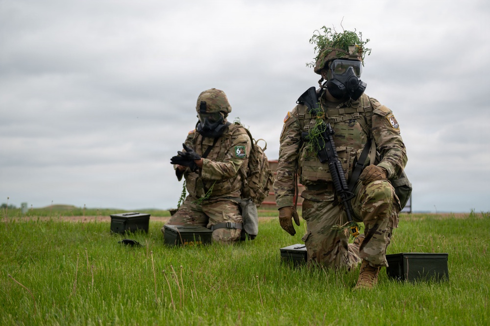Two military personnel in full combat gear, including gas masks and camouflage uniforms, are positioned in an open grassy field. Both soldiers are equipped with tactical vests, weapons, and ammunition cans, and have vegetation attached to their helmets for camouflage. The scene appears to depict a military training exercise conducted under overcast skies.