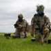 Two military personnel in full combat gear, including gas masks and camouflage uniforms, are positioned in an open grassy field. Both soldiers are equipped with tactical vests, weapons, and ammunition cans, and have vegetation attached to their helmets for camouflage. The scene appears to depict a military training exercise conducted under overcast skies.
