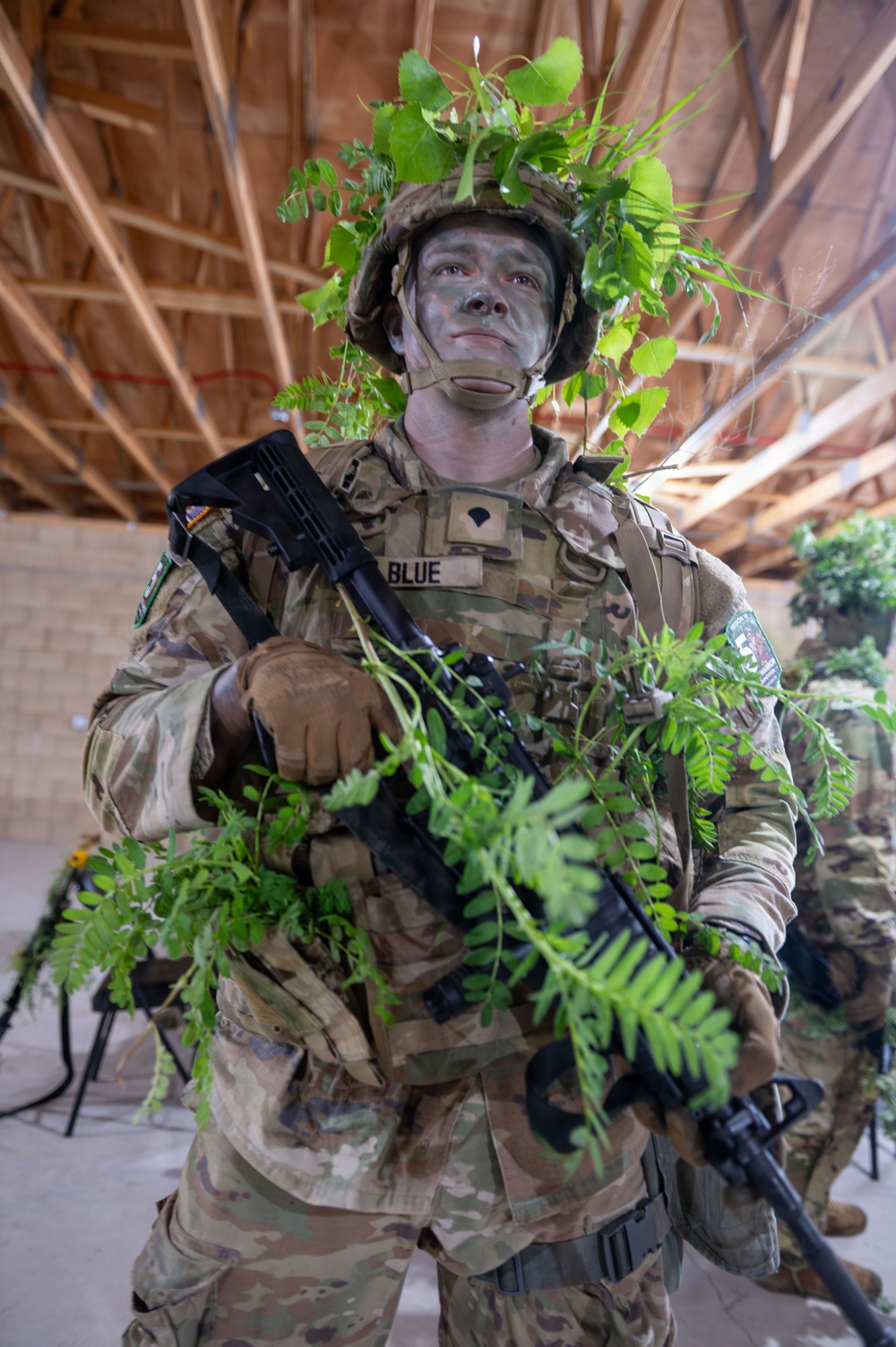 A soldier in full camouflage gear stands inside an unfinished building with exposed wooden rafters. The soldier, identified by a name tape reading 'BLUE,' is wearing MultiCam/OCP pattern uniform with tactical vest, helmet adorned with fresh green foliage, and face paint in green and gray tones. The soldier is holding a black rifle and has additional vegetation attached to their weapon and body for concealment purposes.