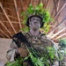 A soldier in full camouflage gear stands inside an unfinished building with exposed wooden rafters. The soldier, identified by a name tape reading 'BLUE,' is wearing MultiCam/OCP pattern uniform with tactical vest, helmet adorned with fresh green foliage, and face paint in green and gray tones. The soldier is holding a black rifle and has additional vegetation attached to their weapon and body for concealment purposes.