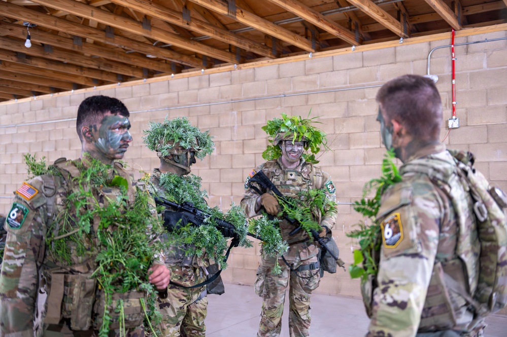 Four military personnel in camouflage uniforms are gathered inside a structure with exposed wooden rafters and a cinder block wall. Three of the soldiers have applied face paint and adorned their helmets and weapons with natural vegetation as camouflage. The group appears to be in a training environment, with at least one soldier smiling, suggesting a relaxed or instructional moment.