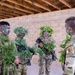 Four military personnel in camouflage uniforms are gathered inside a structure with exposed wooden rafters and a cinder block wall. Three of the soldiers have applied face paint and adorned their helmets and weapons with natural vegetation as camouflage. The group appears to be in a training environment, with at least one soldier smiling, suggesting a relaxed or instructional moment.