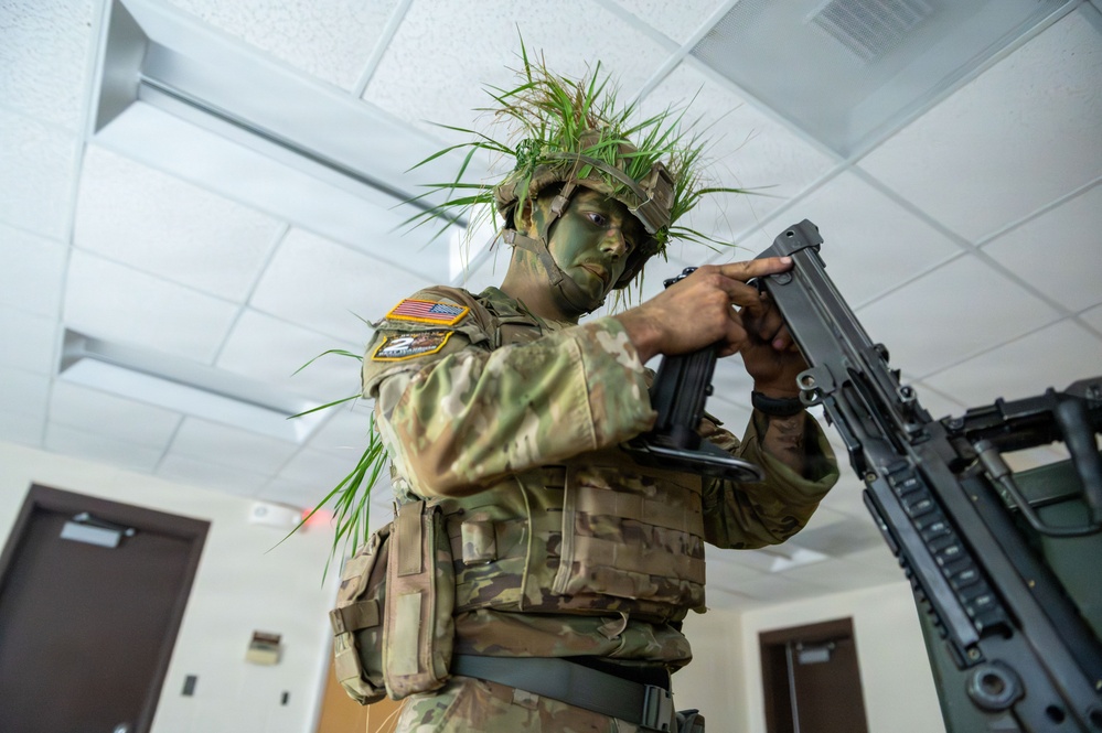 A U.S. military soldier in full camouflage gear, including green face paint and grass/vegetation attached to their helmet, is handling a weapon inside what appears to be an indoor facility. The soldier is wearing MultiCam (OCP) pattern uniform with a U.S. flag patch and additional unit patches visible on the left sleeve. The setting appears to be a training or demonstration environment.