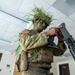 A U.S. military soldier in full camouflage gear, including green face paint and grass/vegetation attached to their helmet, is handling a weapon inside what appears to be an indoor facility. The soldier is wearing MultiCam (OCP) pattern uniform with a U.S. flag patch and additional unit patches visible on the left sleeve. The setting appears to be a training or demonstration environment.
