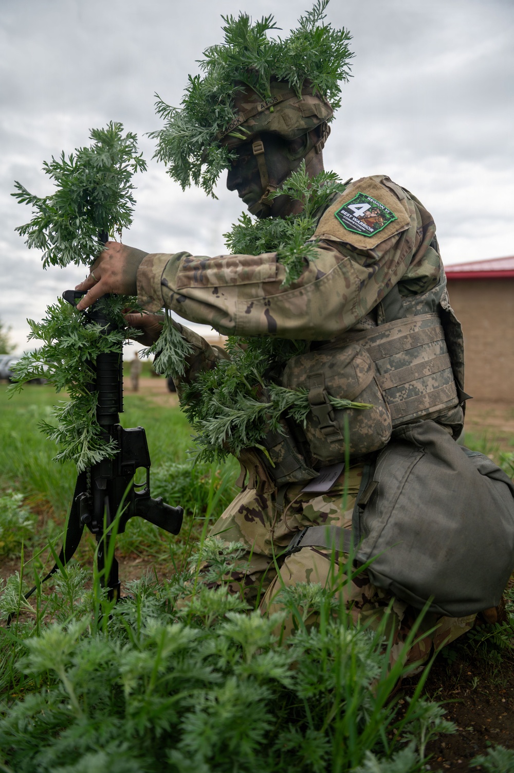 A soldier in full camouflage, including natural vegetation (ghillie-style foliage) attached to their uniform and helmet, kneels in a grassy field while attaching or adjusting foliage to their rifle. The soldier's face is painted dark for concealment, and they wear a 'Region 4 Best Warrior' patch on their left shoulder. This appears to be a military camouflage or stalking competition/training exercise.