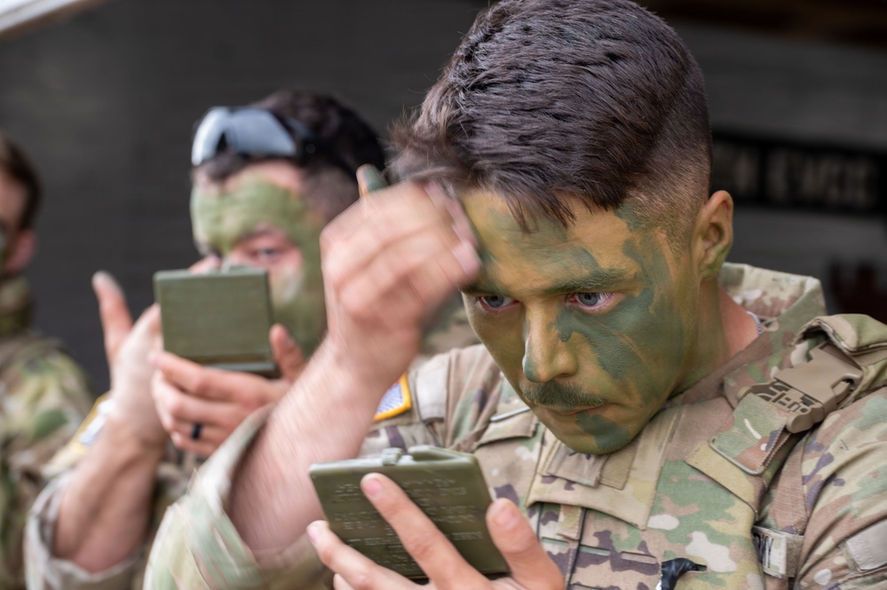 Two military personnel in MultiCam/OCP uniforms are applying camouflage face paint using small compact mirrors. The foreground soldier is carefully applying green and dark camouflage paint to his face, while a second soldier does the same in the background. This appears to be a pre-mission or training preparation activity.