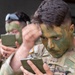 Two military personnel in MultiCam/OCP uniforms are applying camouflage face paint using small compact mirrors. The foreground soldier is carefully applying green and dark camouflage paint to his face, while a second soldier does the same in the background. This appears to be a pre-mission or training preparation activity.