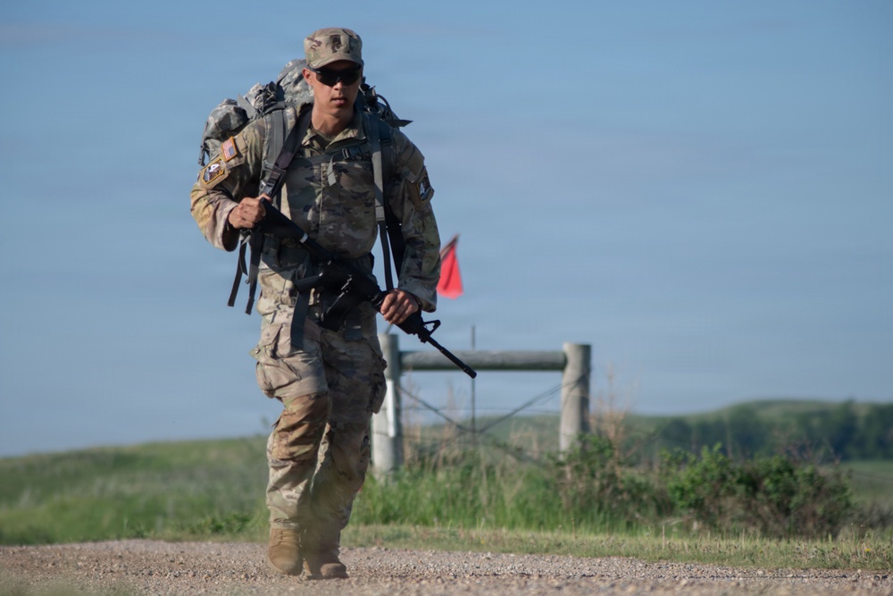A U.S. Army soldier in full MultiCam (OCP) camouflage uniform is conducting a ruck march along a gravel path in an open, grassy terrain. The soldier is carrying a large rucksack on their back and holding a rifle, suggesting a training exercise or physical fitness event. A red marker flag and a fence gate are visible in the background.