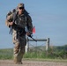 A U.S. Army soldier in full MultiCam (OCP) camouflage uniform is conducting a ruck march along a gravel path in an open, grassy terrain. The soldier is carrying a large rucksack on their back and holding a rifle, suggesting a training exercise or physical fitness event. A red marker flag and a fence gate are visible in the background.