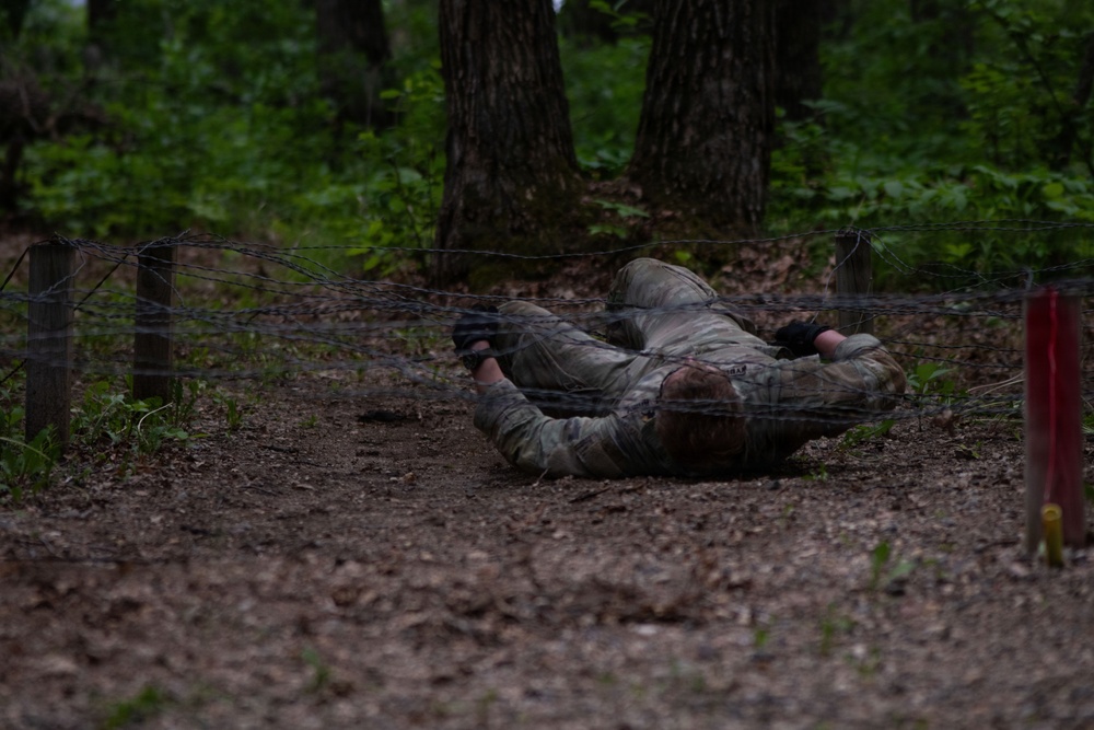 A person in military camouflage uniform is crawling under a low barbed wire obstacle in a wooded training area. The individual is performing a low crawl technique, keeping their body flat against the ground to pass beneath the wire barrier. This appears to be a military or tactical obstacle course training exercise.