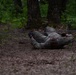 A person in military camouflage uniform is crawling under a low barbed wire obstacle in a wooded training area. The individual is performing a low crawl technique, keeping their body flat against the ground to pass beneath the wire barrier. This appears to be a military or tactical obstacle course training exercise.