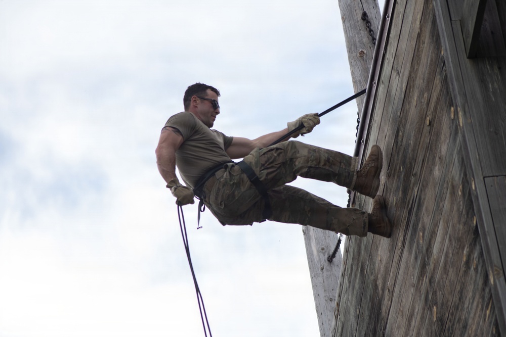 A male individual in military-style clothing is rappelling down a large wooden wall structure. He is suspended by ropes with a harness system, with his feet braced against the wooden surface in a classic rappelling posture. The image is taken from a low angle looking upward, emphasizing the height and physical effort involved.