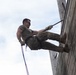 A male individual in military-style clothing is rappelling down a large wooden wall structure. He is suspended by ropes with a harness system, with his feet braced against the wooden surface in a classic rappelling posture. The image is taken from a low angle looking upward, emphasizing the height and physical effort involved.