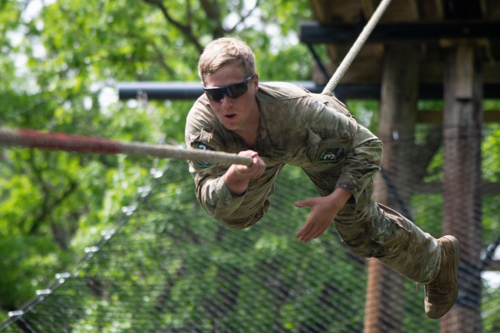 A soldier in military camouflage uniform is swinging on a rope during what appears to be a military obstacle course or training exercise. The individual is airborne, gripping a horizontal rope with both hands while their body is suspended mid-swing. The setting includes wooden platform structures and netting consistent with a military training facility.