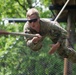 A soldier in military camouflage uniform is swinging on a rope during what appears to be a military obstacle course or training exercise. The individual is airborne, gripping a horizontal rope with both hands while their body is suspended mid-swing. The setting includes wooden platform structures and netting consistent with a military training facility.