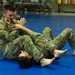 Two military personnel in OCP (Operational Camouflage Pattern) uniforms are practicing ground combat/grappling techniques on a blue training mat. One soldier is in a dominant top position while the other is on their back, with both engaging in what appears to be a controlled combatives training drill. Additional uniformed personnel are visible observing in the background.