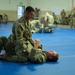 Military personnel are engaged in combatives (hand-to-hand combat) training on blue mats inside a gymnasium or training facility. In the foreground, one soldier in camouflage uniform has mounted another soldier who is on his back, simulating a ground fighting technique. Multiple other pairs of soldiers are practicing similar techniques in the background.