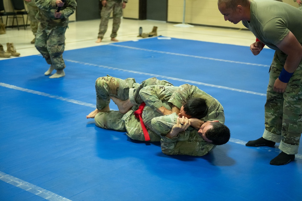 Military personnel in camouflage uniforms are engaged in a combatives/grappling training session on blue mats in an indoor facility. Two soldiers are grappling on the ground while a referee or instructor leans in to observe closely. Additional uniformed personnel are visible in the background observing the match.