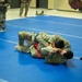 Military personnel in camouflage uniforms are engaged in a combatives/grappling training session on blue mats in an indoor facility. Two soldiers are grappling on the ground while a referee or instructor leans in to observe closely. Additional uniformed personnel are visible in the background observing the match.