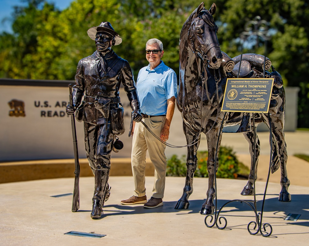 A man in a light blue polo shirt and khaki pants stands between two bronze sculptures at what appears to be a U.S. Army installation. The sculptures depict a Buffalo Soldier leading a horse, with a plaque identifying the memorial as honoring Congressional Medal of Honor Recipient William H. Thompkins. The setting is an outdoor memorial plaza with a wall bearing the text 'U.S. Army... Read... A...' partially visible in the background.