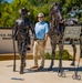 A man in a light blue polo shirt and khaki pants stands between two bronze sculptures at what appears to be a U.S. Army installation. The sculptures depict a Buffalo Soldier leading a horse, with a plaque identifying the memorial as honoring Congressional Medal of Honor Recipient William H. Thompkins. The setting is an outdoor memorial plaza with a wall bearing the text 'U.S. Army... Read... A...' partially visible in the background.