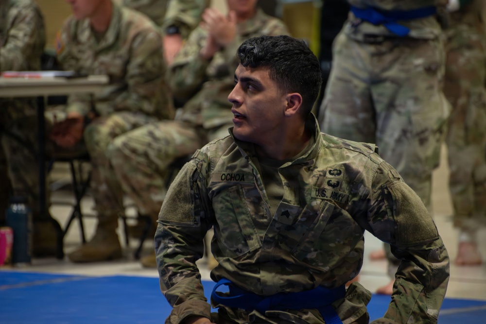 A U.S. Army soldier named Ochoa is kneeling on a blue martial arts mat, wearing an OCP (Operational Camouflage Pattern) uniform with a blue belt, indicating participation in a combatives competition or training event. He appears focused and attentive, looking off to the side. Other soldiers in uniform are visible in the blurred background, some seated at tables, suggesting an organized military combatives event.