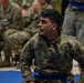 A U.S. Army soldier named Ochoa is kneeling on a blue martial arts mat, wearing an OCP (Operational Camouflage Pattern) uniform with a blue belt, indicating participation in a combatives competition or training event. He appears focused and attentive, looking off to the side. Other soldiers in uniform are visible in the blurred background, some seated at tables, suggesting an organized military combatives event.