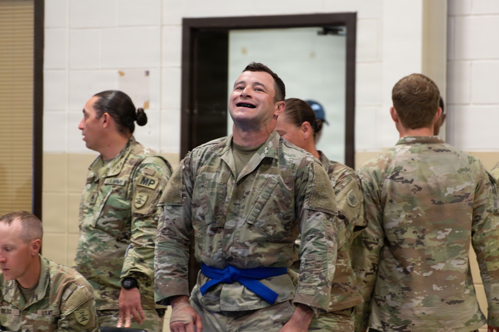 A group of U.S. Army soldiers in Operational Camouflage Pattern (OCP) uniforms are gathered in an indoor facility. The central figure is laughing or reacting expressively with his head tilted back, wearing a blue belt around his uniform, suggesting a combatives or martial arts training context. Other soldiers are visible in the background and foreground, some with 'MP' (Military Police) patches.