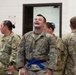 A group of U.S. Army soldiers in Operational Camouflage Pattern (OCP) uniforms are gathered in an indoor facility. The central figure is laughing or reacting expressively with his head tilted back, wearing a blue belt around his uniform, suggesting a combatives or martial arts training context. Other soldiers are visible in the background and foreground, some with 'MP' (Military Police) patches.