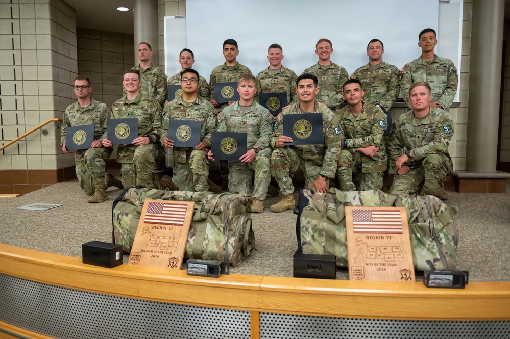 A group of approximately 14 U.S. Army soldiers in OCP (Operational Camouflage Pattern) uniforms pose for a formal group photo. Several soldiers in the front row are holding dark certificate folders with gold seals. In the foreground, two wooden plaques and military duffel bags are displayed, with the plaques reading 'Region VI Soldier of the Year 2026' and 'Region VI NCO of the Year 2026' for the Army National Guard.