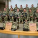 A group of approximately 14 U.S. Army soldiers in OCP (Operational Camouflage Pattern) uniforms pose for a formal group photo. Several soldiers in the front row are holding dark certificate folders with gold seals. In the foreground, two wooden plaques and military duffel bags are displayed, with the plaques reading 'Region VI Soldier of the Year 2026' and 'Region VI NCO of the Year 2026' for the Army National Guard.