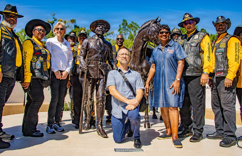 A group of people pose around a bronze memorial statue depicting a Buffalo Soldier standing with a rifle and a horse. Several individuals are wearing distinctive black leather vests with yellow shirts bearing patches that read 'NABS MC' and 'Buffalo Soldiers,' indicating membership in a Buffalo Soldiers motorcycle club. One man kneels in the foreground in front of the statue, suggesting this may be a dedication or commemorative event.