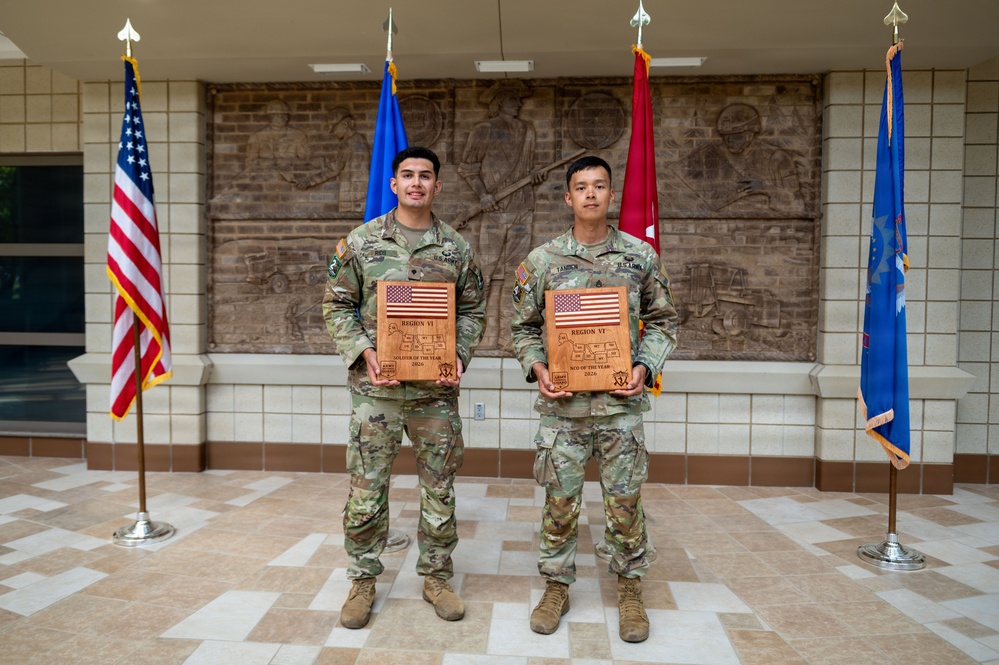 Two U.S. Army soldiers in OCP (Operational Camouflage Pattern) uniforms stand side by side holding wooden plaques in what appears to be a military building lobby. The plaques read 'Region VI' and indicate awards for 'Soldier of the Year 2024' and 'NCO of the Year 2024' respectively. The formal setting with multiple flags suggests this is an official awards ceremony or recognition photo.