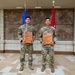 Two U.S. Army soldiers in OCP (Operational Camouflage Pattern) uniforms stand side by side holding wooden plaques in what appears to be a military building lobby. The plaques read 'Region VI' and indicate awards for 'Soldier of the Year 2024' and 'NCO of the Year 2024' respectively. The formal setting with multiple flags suggests this is an official awards ceremony or recognition photo.
