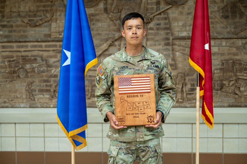 A U.S. Army soldier named Tangen stands at attention holding a wooden award plaque. The plaque reads 'Region VI NCO of the Year 2026' and features an engraved American flag and a map of northwestern U.S. states along with Army National Guard insignia. The soldier is flanked by two ceremonial flags in what appears to be a formal military building interior.