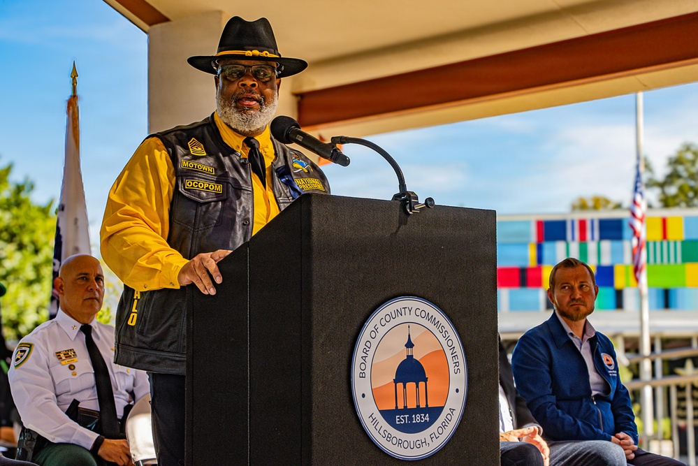 A man wearing a black leather vest with patches, a yellow shirt, and a black wide-brimmed hat is speaking at a podium bearing the seal of the Board of County Commissioners, Hillsborough, Florida (Est. 1834). The event appears to be an official outdoor ceremony, with two other individuals seated in the background. Colorful panels, an American flag, and what appears to be a military or ceremonial color guard flag are visible in the background.