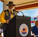 A man wearing a black leather vest with patches, a yellow shirt, and a black wide-brimmed hat is speaking at a podium bearing the seal of the Board of County Commissioners, Hillsborough, Florida (Est. 1834). The event appears to be an official outdoor ceremony, with two other individuals seated in the background. Colorful panels, an American flag, and what appears to be a military or ceremonial color guard flag are visible in the background.