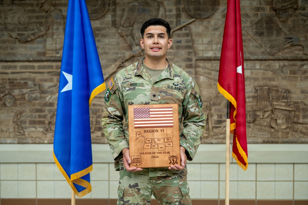 A U.S. Army National Guard soldier in OCP (Operational Camouflage Pattern) uniform holds a wooden award plaque recognizing him as the Region VI Soldier of the Year 2026. He is standing between two flags — a blue flag with a star and a red general officer flag — in front of a brick wall with military relief sculptures. His name tape reads 'RIOS' and he is wearing U.S. Army insignia.