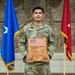 A U.S. Army National Guard soldier in OCP (Operational Camouflage Pattern) uniform holds a wooden award plaque recognizing him as the Region VI Soldier of the Year 2026. He is standing between two flags — a blue flag with a star and a red general officer flag — in front of a brick wall with military relief sculptures. His name tape reads 'RIOS' and he is wearing U.S. Army insignia.