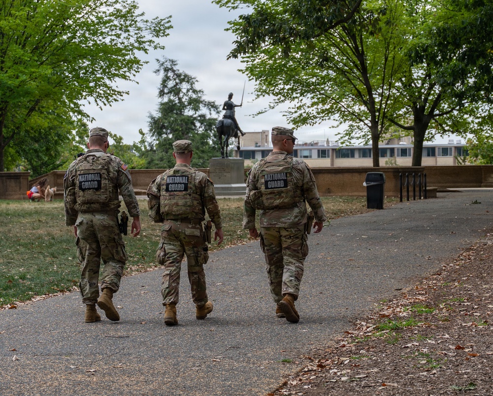 Three National Guard soldiers in full camouflage uniforms and tactical vests are walking away from the camera along a paved park path. A bronze equestrian statue is visible in the background amid green trees. The scene appears to be a patrol or security detail in an urban park setting.