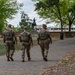 Three National Guard soldiers in full camouflage uniforms and tactical vests are walking away from the camera along a paved park path. A bronze equestrian statue is visible in the background amid green trees. The scene appears to be a patrol or security detail in an urban park setting.