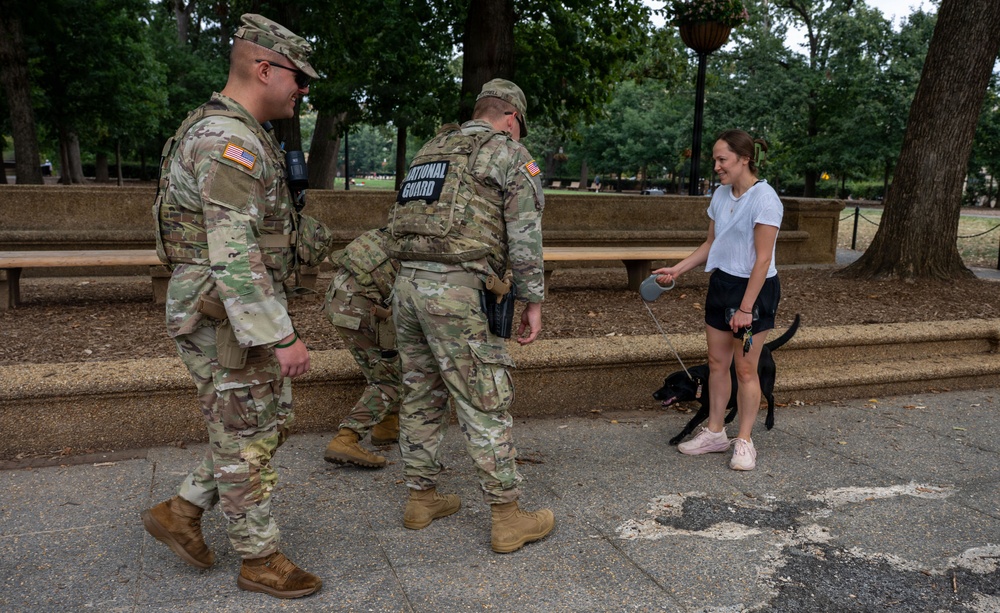Two U.S. National Guard soldiers in OCP (Operational Camouflage Pattern) uniforms interact with a civilian woman walking a black dog in what appears to be a public park. The soldiers are in full tactical gear including body armor, and one has a clearly visible 'National Guard' patch on their back. The setting appears to be an urban park with benches, trees, and paved walkways.
