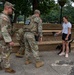 Two U.S. National Guard soldiers in OCP (Operational Camouflage Pattern) uniforms interact with a civilian woman walking a black dog in what appears to be a public park. The soldiers are in full tactical gear including body armor, and one has a clearly visible 'National Guard' patch on their back. The setting appears to be an urban park with benches, trees, and paved walkways.