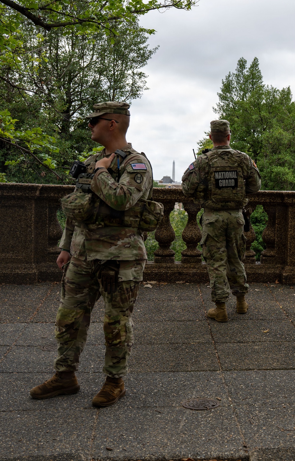 Two U.S. National Guard soldiers in full combat gear are standing on a stone terrace or balustrade, appearing to conduct a security patrol or observation post. Both wear OCP (Operational Camouflage Pattern) uniforms with American flag patches and tactical vests. In the background, a tall obelisk-shaped monument — consistent with the Washington Monument — is visible through the trees, suggesting this location is in Washington, D.C.
